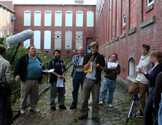 An outdoor audio drama cast and crew recording with a boom microphone beside historic brick buildings.